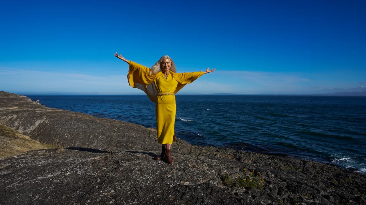 Iana Lahi in gold on the ocean rocks, arms outstretched